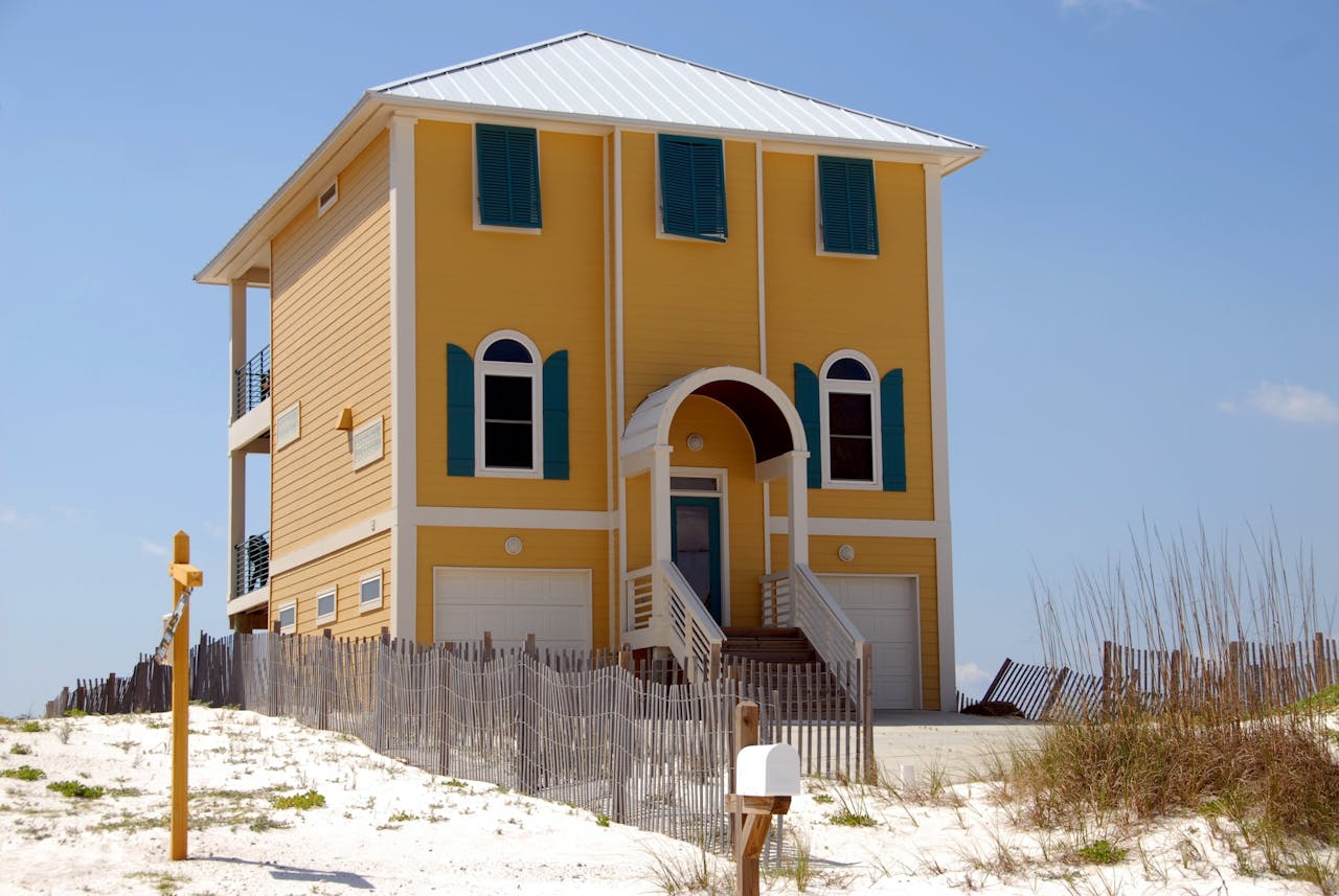 why-choose-us A vibrant yellow beach house with green shutters sits on a sandy dune, surrounded by a fence.